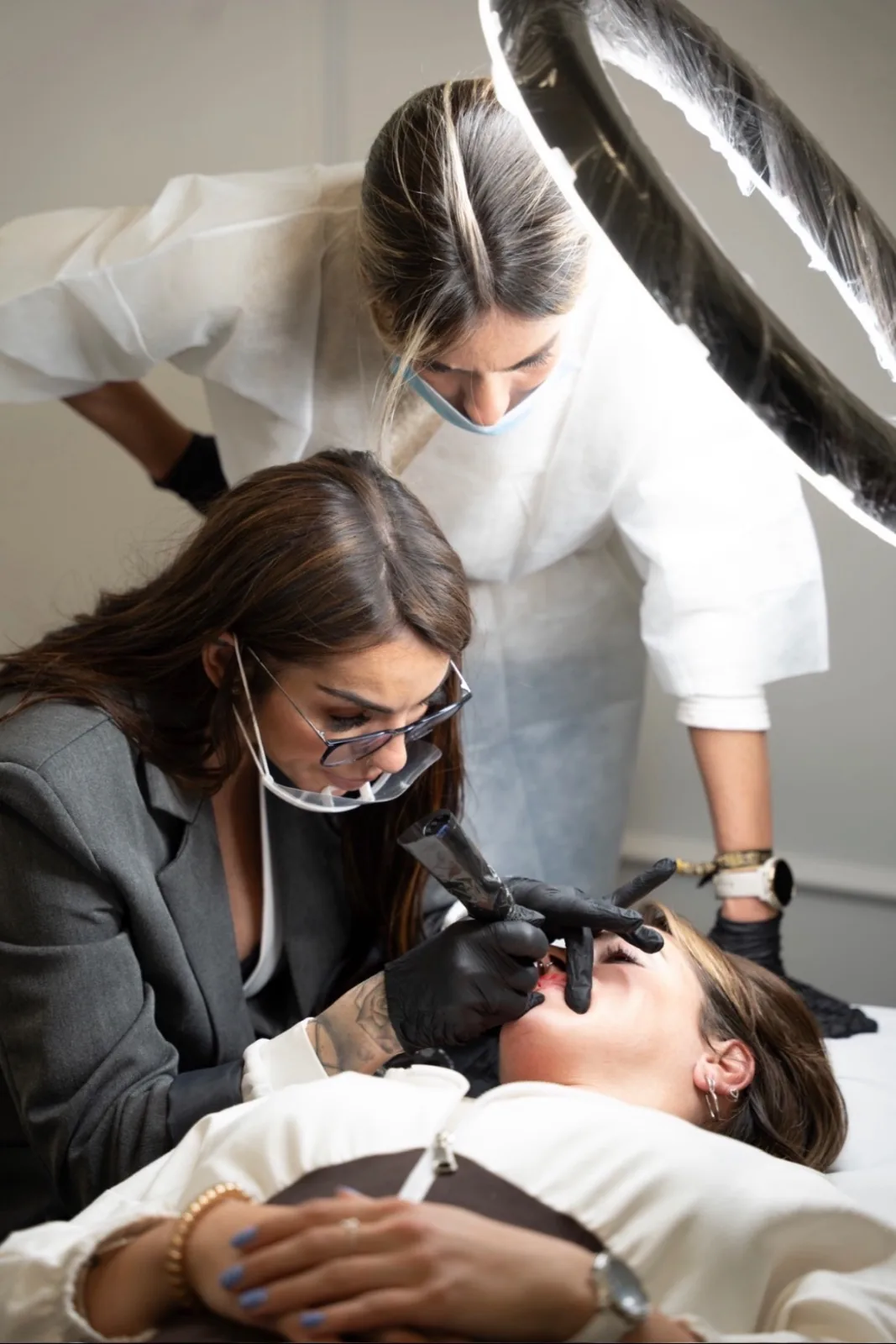 Esteticista con mascarilla y guantes realizando micropigmentación de labios con otra profesional supervisando