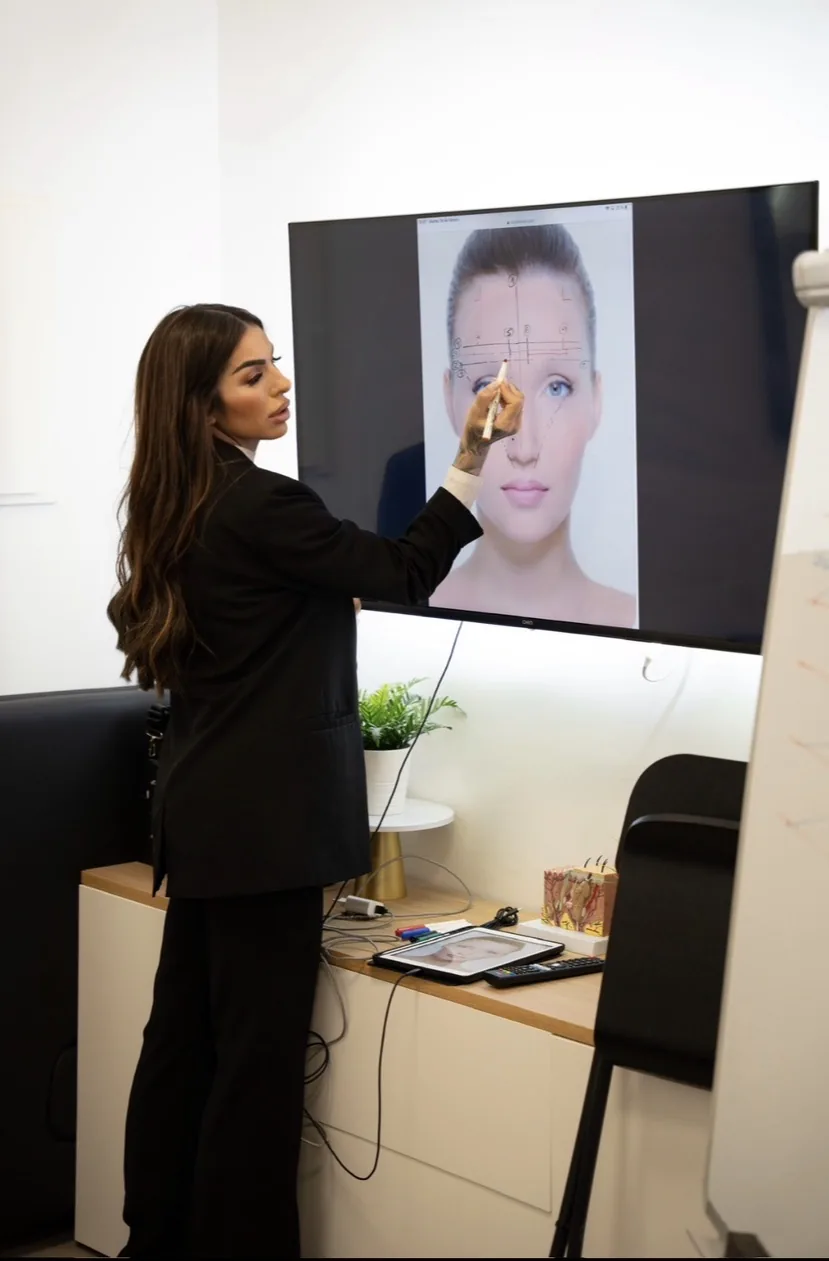Formadora marcando con un lápiz las medidas de cejas sobre una pantalla en el aula