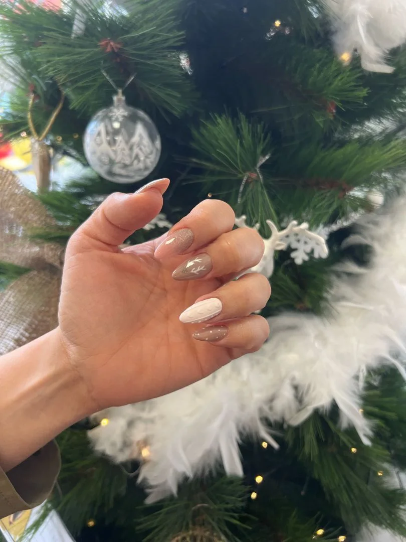 Mano con uñas almendradas nude y blancas con motivos navideños frente a un árbol