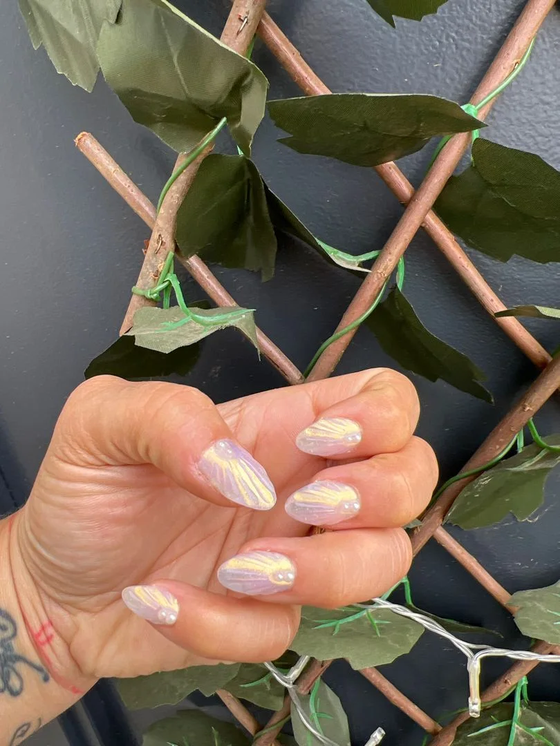 Mano con uñas almendradas efecto concha nacarada irisada sobre fondo de hojas verdes