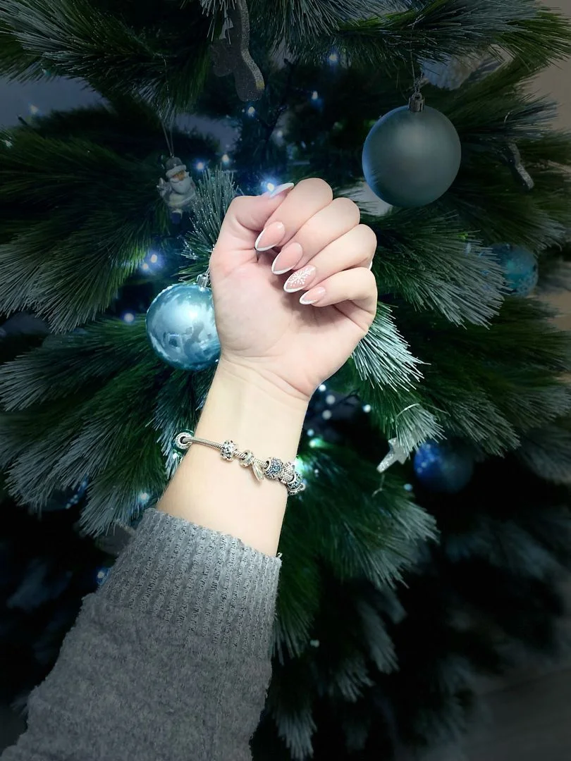 Mano con uñas almendradas nude y diseño de copo de nieve junto a árbol de Navidad